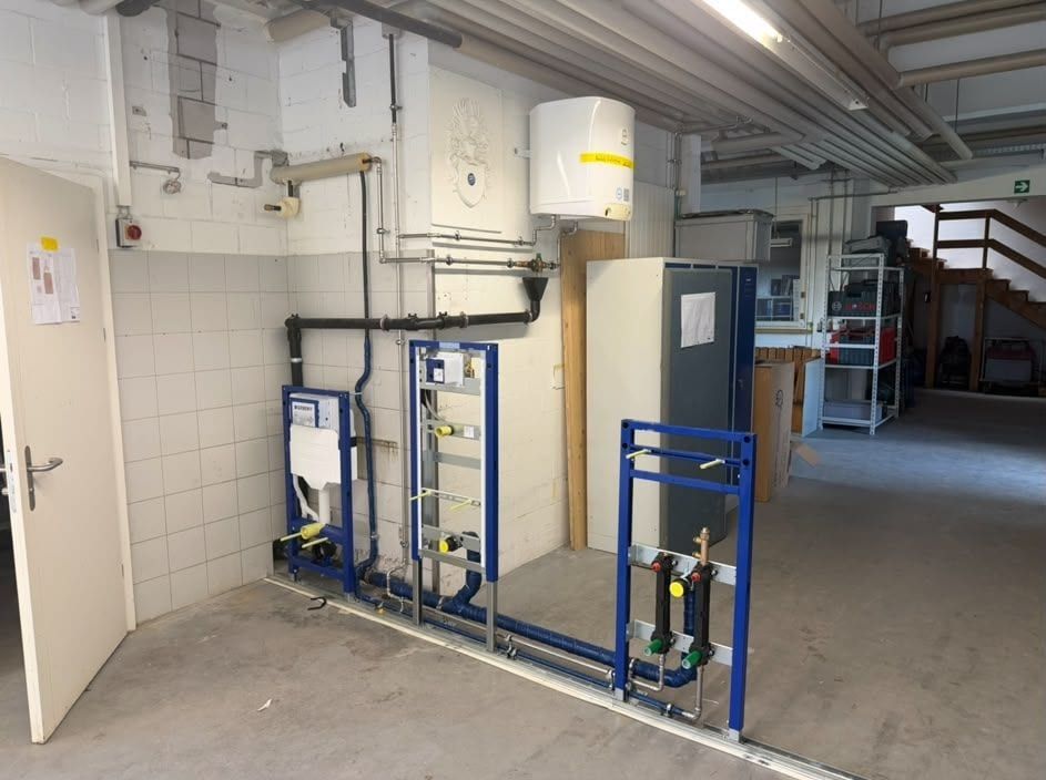Blaue Geberit-Vorwandelemente mit Anschlüssen vor einer gefliesten Wand, darüber ein Boiler, am Boden die Leitungen zu den Elementen
