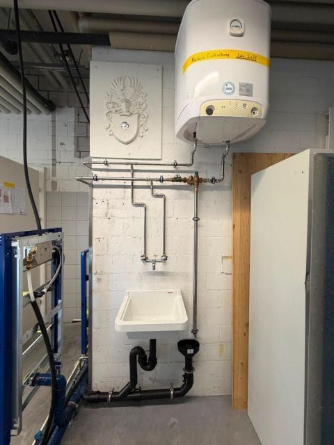 Wandhängender Boiler über einem Ausgussbecken mit Leitungen, links ein blaues Vorwandelement