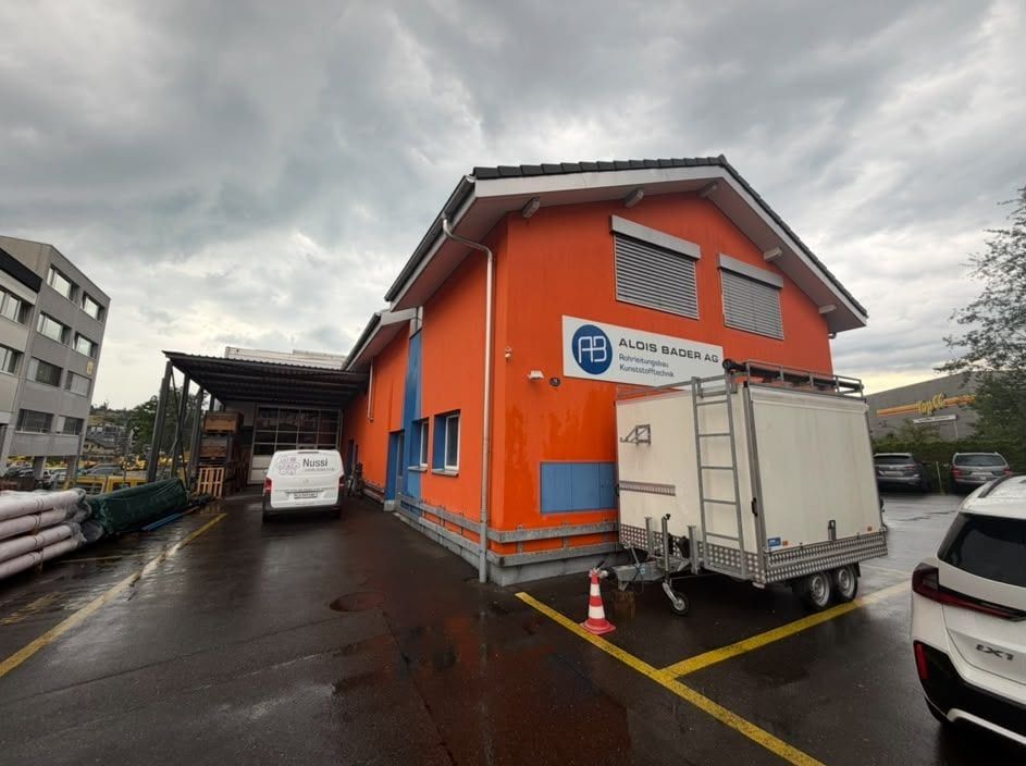 Orangefarbenes Gewerbegebäude mit dem Firmenschild «Alois Bader AG, Rohrleitungsbau, Kunststofftechnik», davor ein Lieferwagen und ein Anhänger