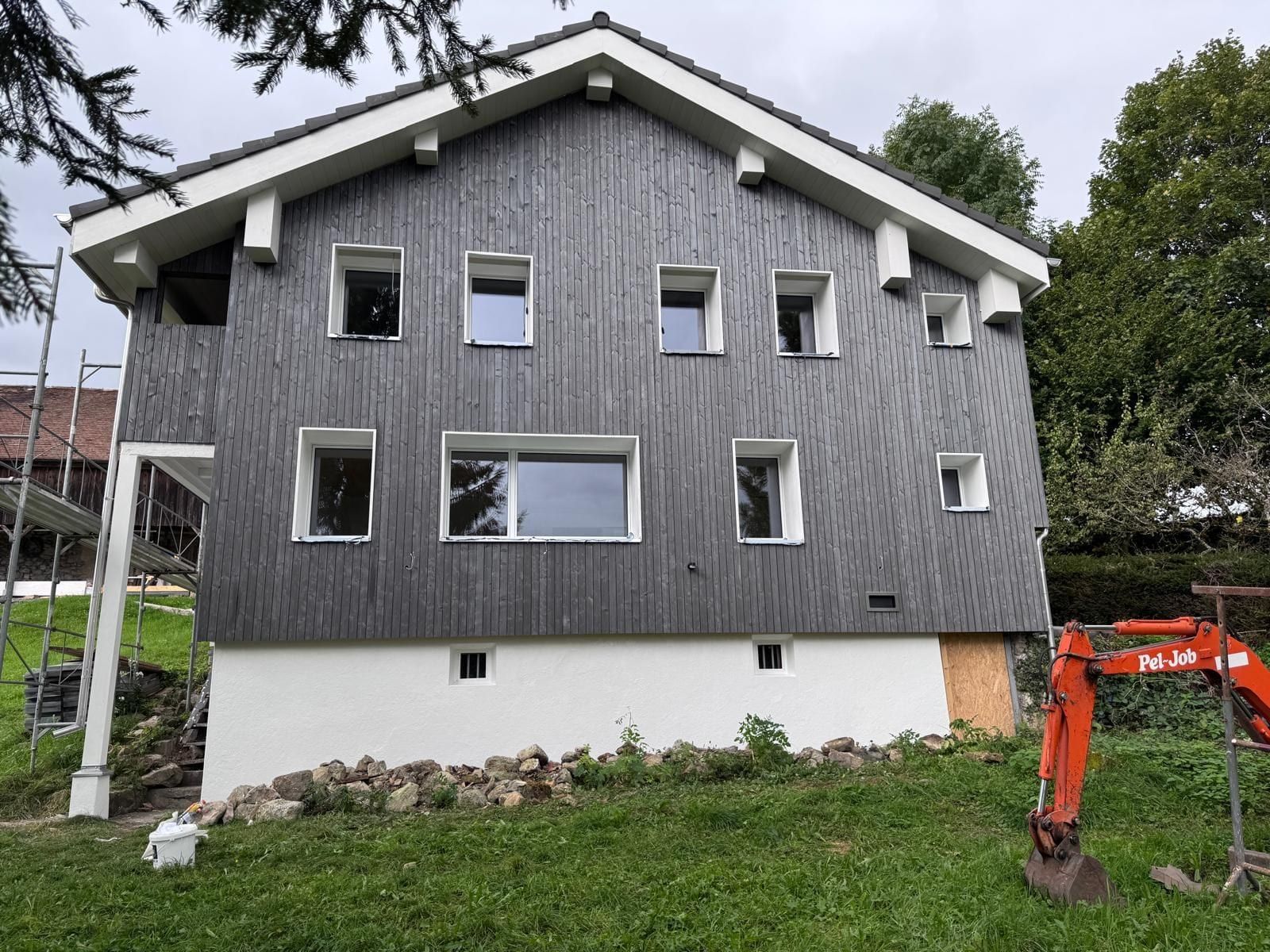 Haus mit dunkler Holzschalung, weissen Fensterumrandungen und weiss verputztem Sockel, links ein Gerüst, rechts ein Kleinbagger
