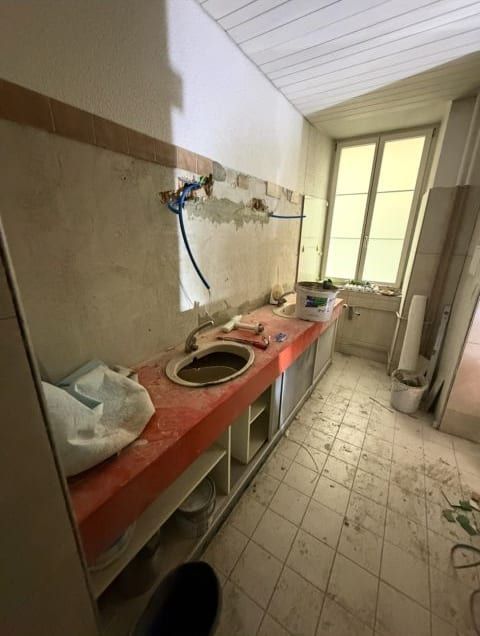 Langer Raum im alten Zustand: rote Abdeckung mit eingebautem Lavabo, offene Leitungen an der Wand, am Ende ein Fenster