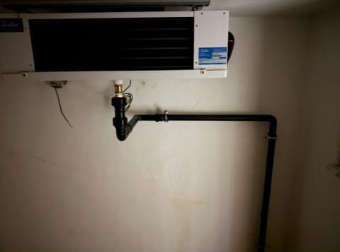 Schwarze Ablaufleitung mit Siphon an einer hellen Wand, darüber ein an der Decke montiertes Gerät