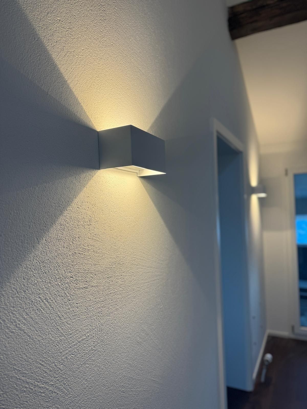 Wand mit Strukturputz, eine würfelförmige Wandleuchte wirft Licht nach oben und unten, im Hintergrund ein Gang